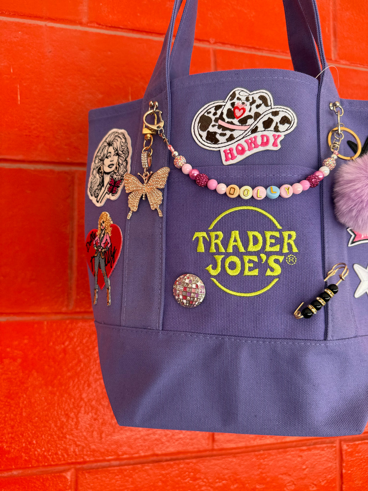 A purple canvas tote bag with 'Trader Joe's' embroidered in yellow on the front, decorated with various patches, a beaded charm, and a fuzzy pom-pom, set against a bright orange brick wall. Dolly Parton Sweet but Strong mini tote by PrettyMUCH.