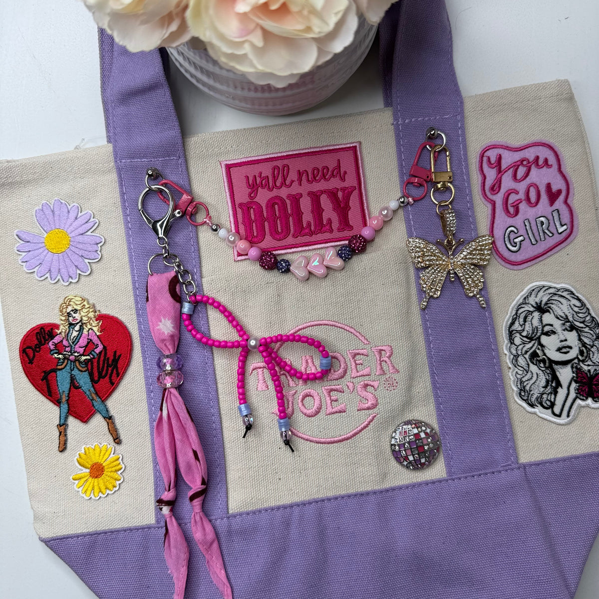 A lavender and cream Trader Joe's mini tote styled with pink and purple accessories, including a pink beaded bag chain, a butterfly charm, a rhinestone disco ball pin and embroidered patches, on a white background with a flower pot. Dolly Parton You Go Girl mini tote by PrettyMUCH.