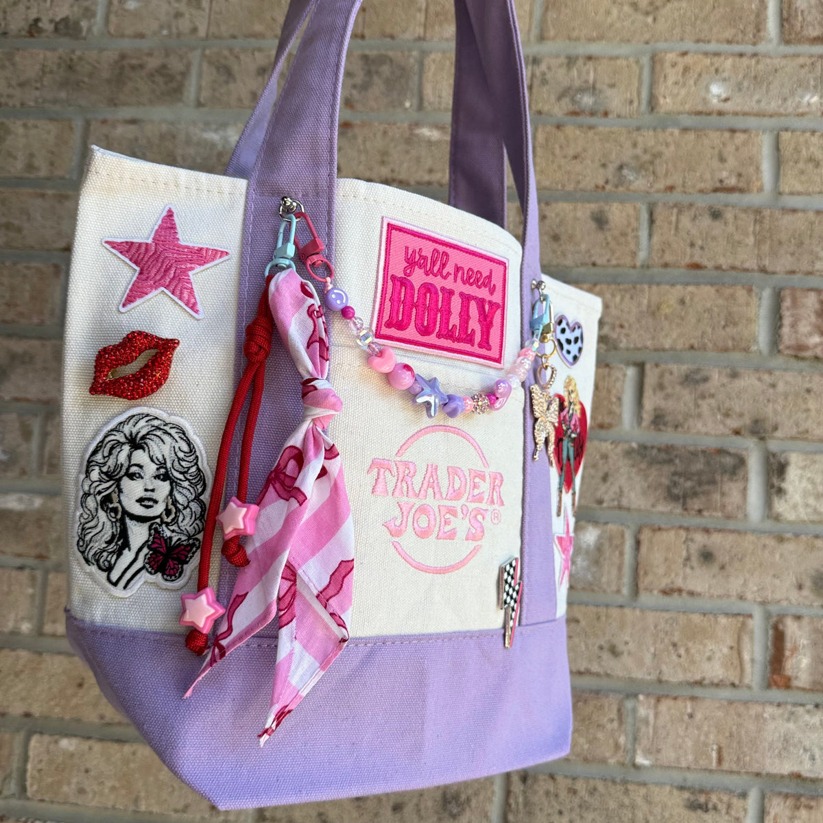 A lavender and cream Trader Joe's mini tote decorated with a hot pink 'y'all need DOLLY' patch, the Trader Joe's logo, pink zebra-stripe star patches and a red rhinestone lips pin, hanging against a brick wall. Dolly Parton Pretty in Pastel mini tote by PrettyMUCH.