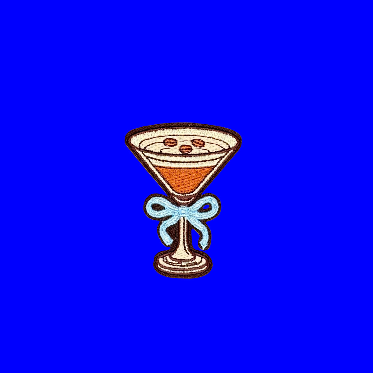 An embroidered patch shaped like a martini glass filled with an espresso martini and espresso beans, tied with a light blue bow, centered on a bright blue background