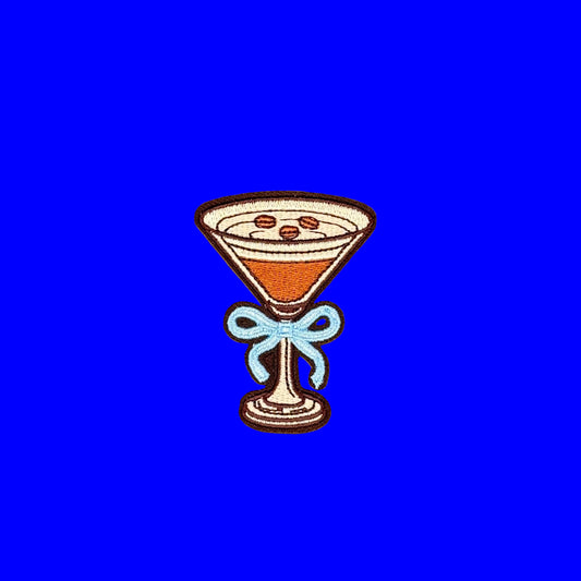 An embroidered patch shaped like a martini glass filled with an espresso martini and espresso beans, tied with a light blue bow, centered on a bright blue background
