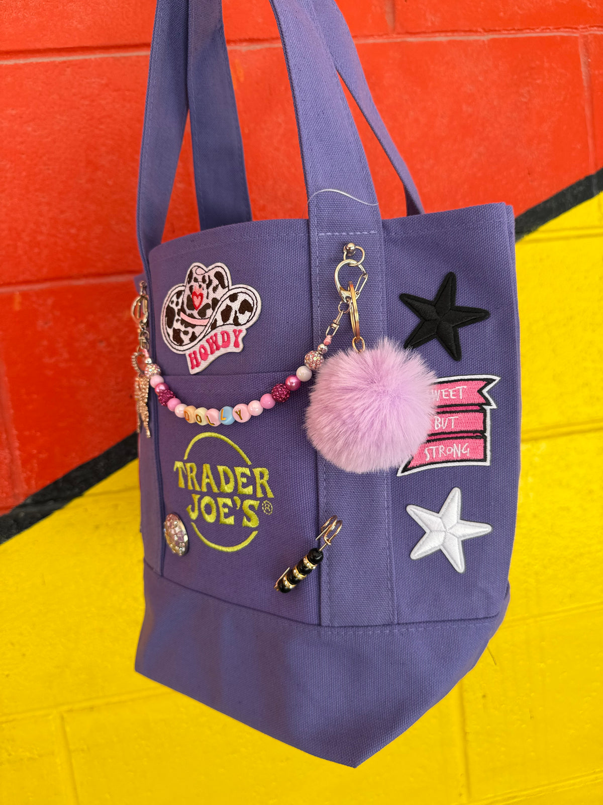 A purple Trader Joe's mini tote with Dolly patches, a lavender pom-pom and a beaded DOLLY bag chain, shown against a bright yellow and orange background. Dolly Parton Sweet but Strong mini tote by PrettyMUCH.