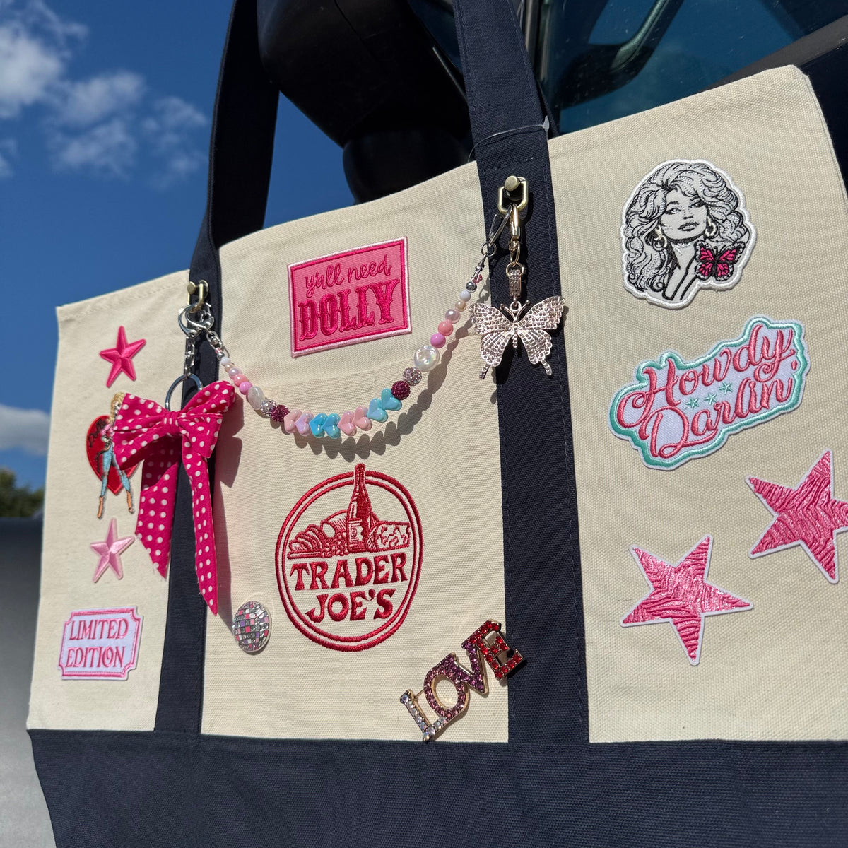 A natural canvas Trader Joe's large tote with navy straps, decorated with Dolly patches, pins and a beaded bag chain, shown outdoors against a blue sky. Dolly Parton Big Love large tote by PrettyMUCH.