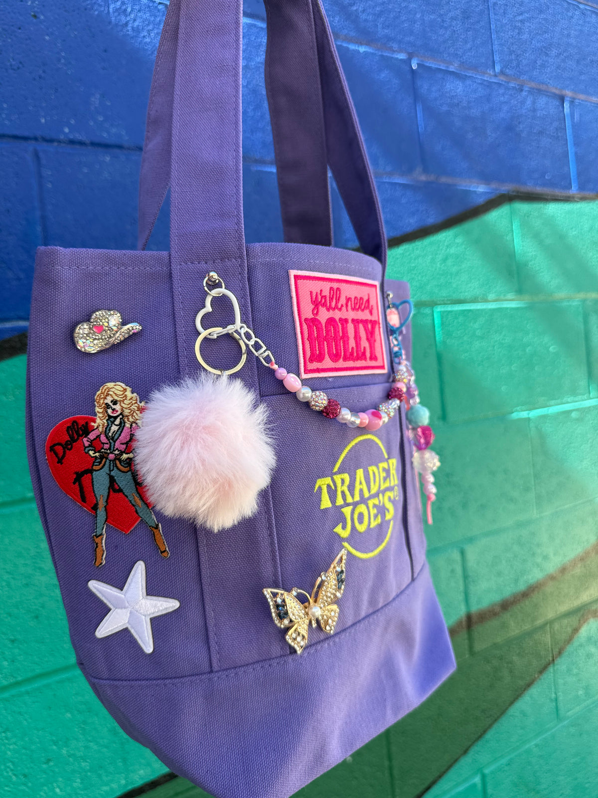 A purple canvas tote bag decorated with multiple patches, pins, a fluffy pom-pom charm, and a beaded keychain, hanging against a blue and green painted brick wall. Dolly Parton Aces High mini tote by PrettyMUCH.