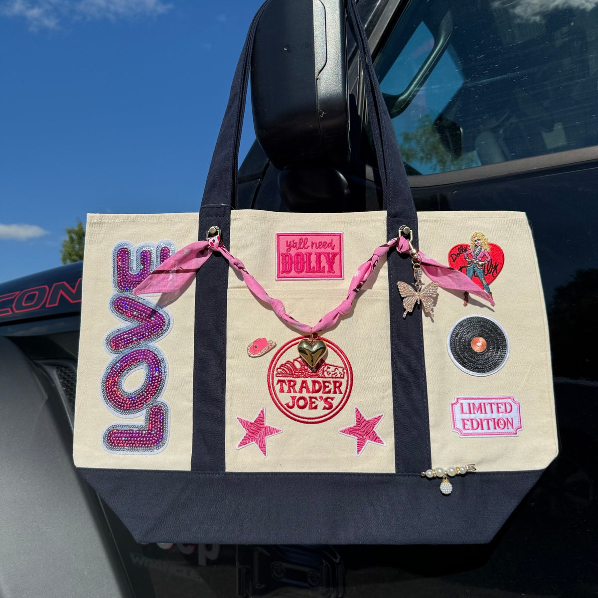 Dolly Parton Limited Edition large tote hanging from a car mirror, showing the pink sequin LOVE patch, y'all need DOLLY patch and vinyl record patch