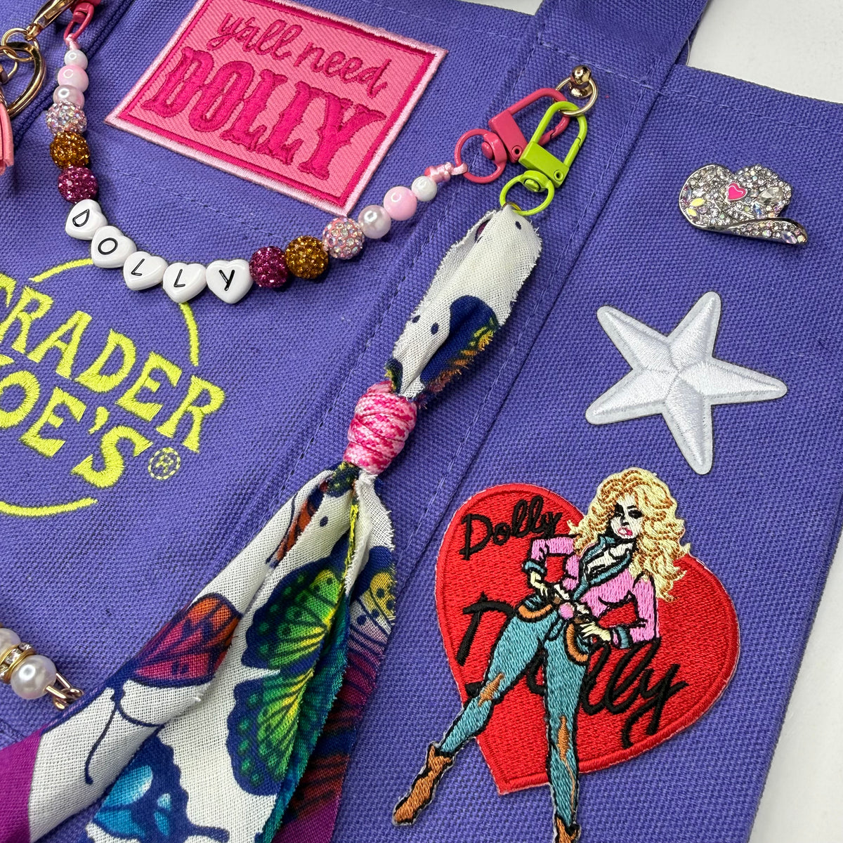 A close-up flat lay of a purple fabric accessory, likely a bag or pouch, featuring a pink 'DOLLY' patch, a beaded letter keychain spelling 'DOLLY', a patterned fabric charm with a tassel, a white star-shaped embellishment, a rhinestone heart pin, and a red heart patch with an illustrated Dolly character. Dolly Parton Y'all Need Dolly mini tote by PrettyMUCH.