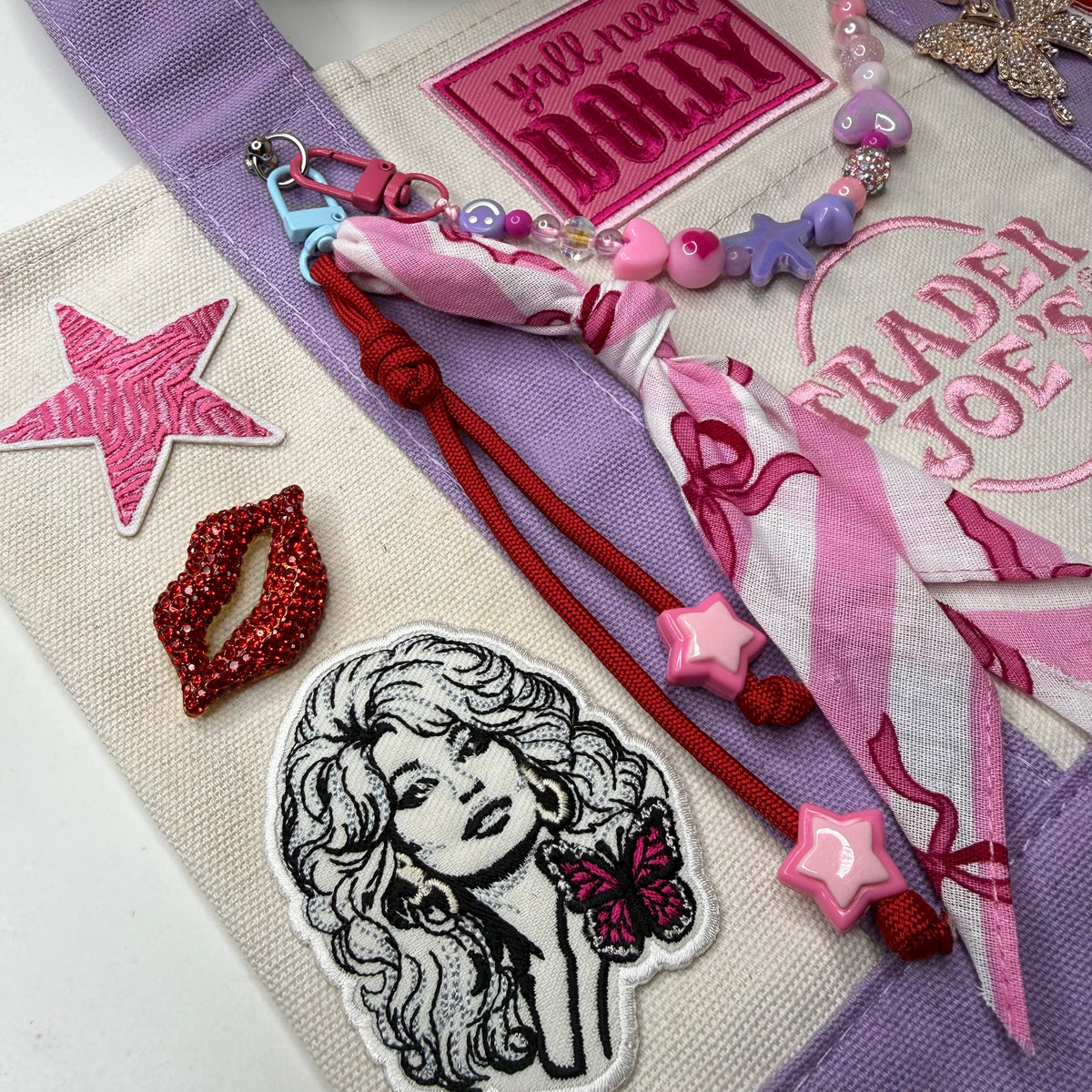 Close-up of the lavender and cream Trader Joe's mini tote with a pink zebra-stripe star patch, a red rhinestone lips pin, a Dolly Parton patch, a pink striped bandana twilly and a beaded bag chain. Dolly Parton Pretty in Pastel mini tote by PrettyMUCH.