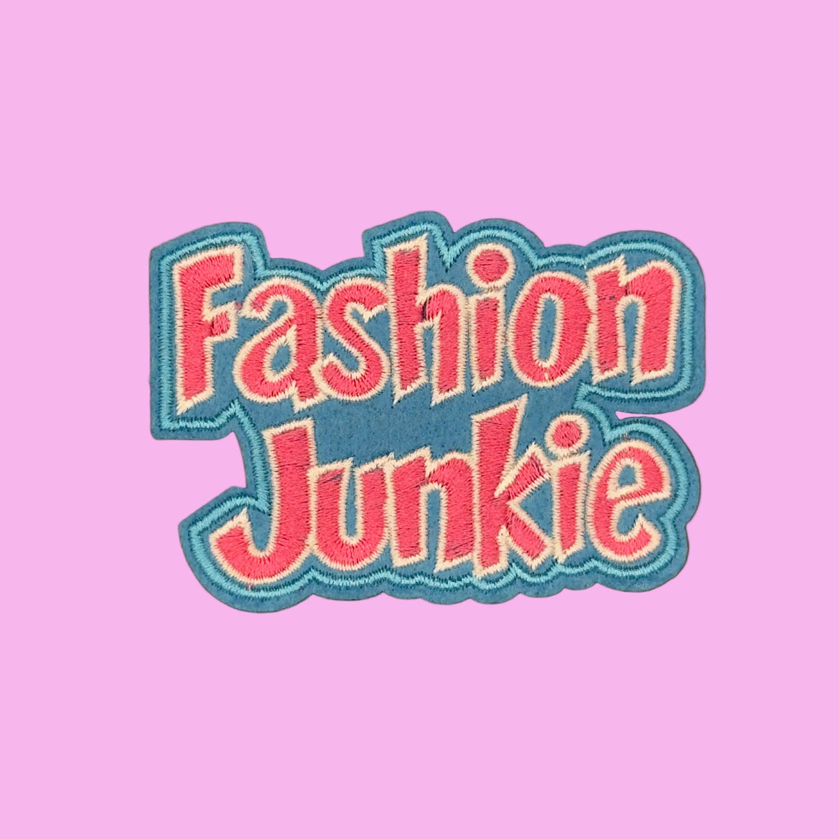 Fashion Junkie in pink letters with a white outline on a blue border embroidered iron-on patch. 