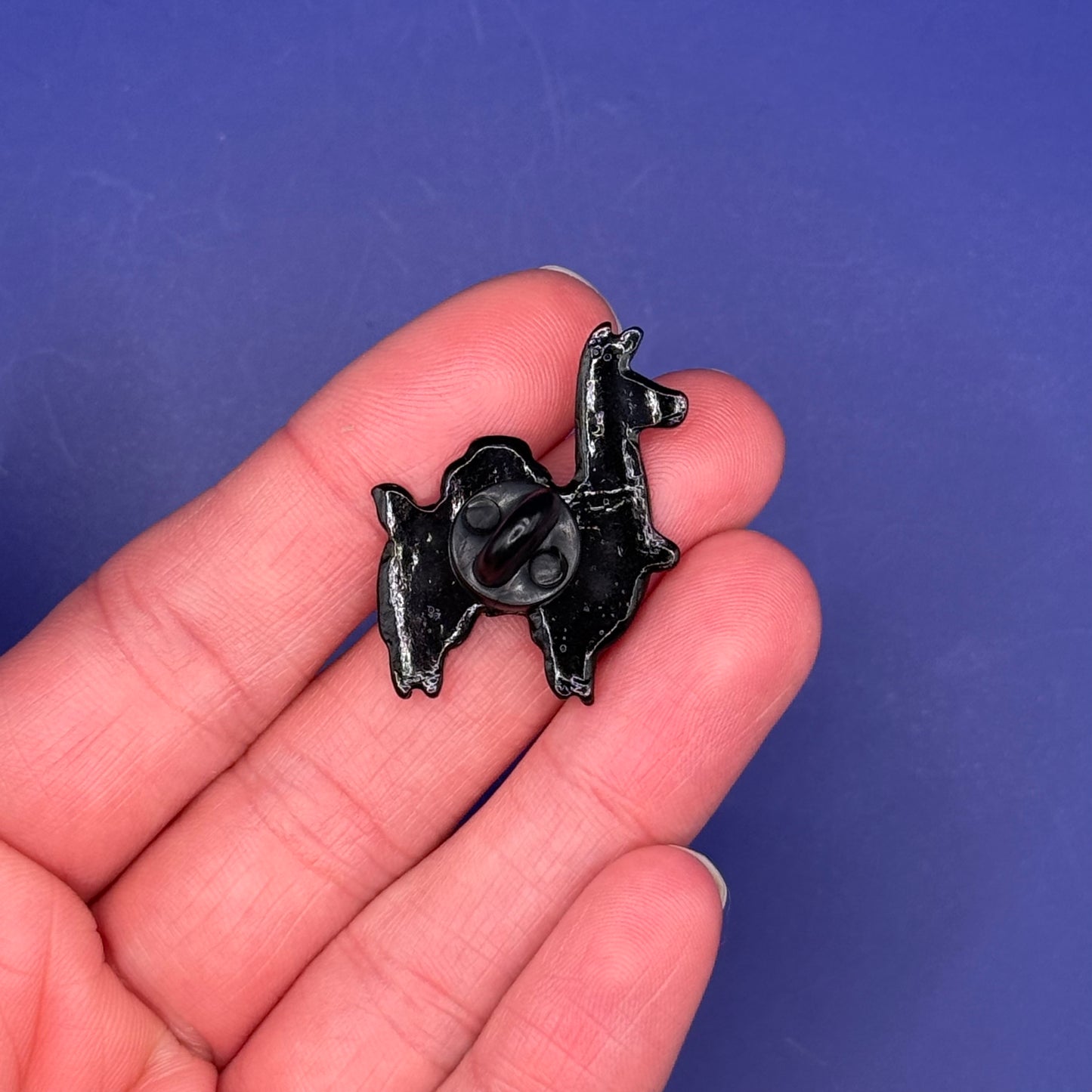 Back of the llama with pastel saddle bag enamel pin showing the black metal back, held in hand