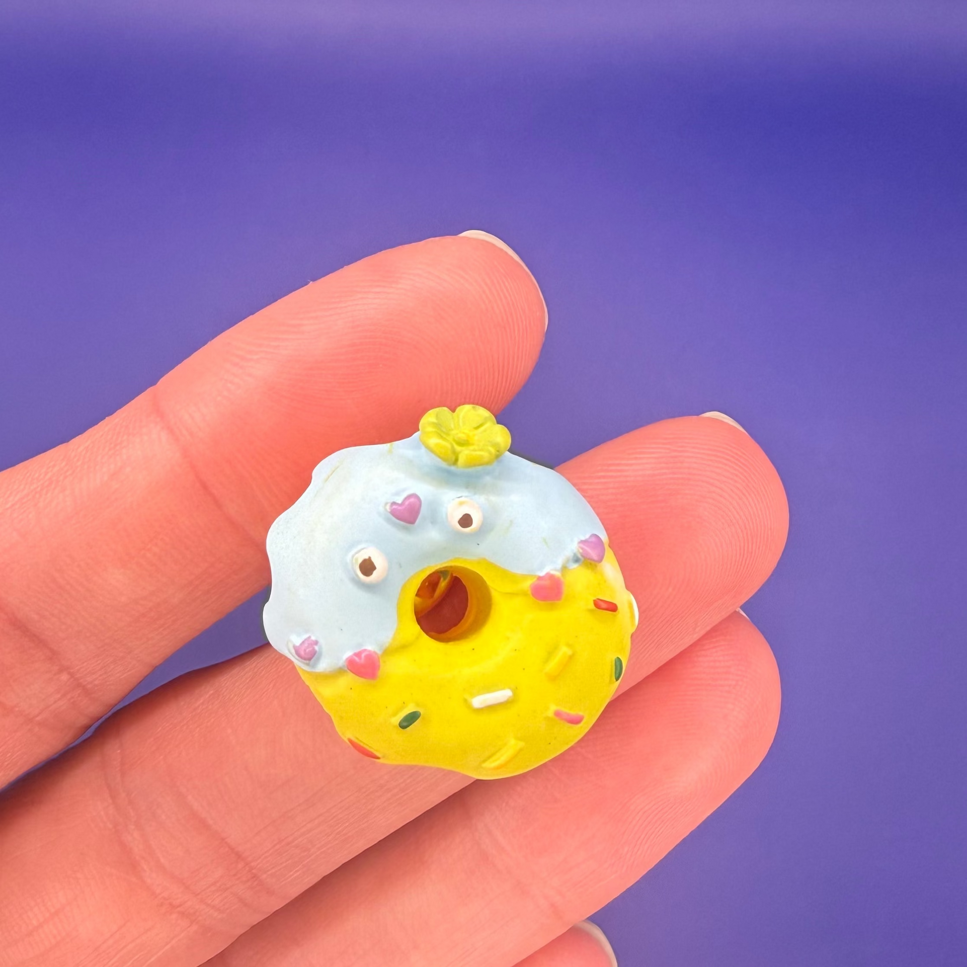 Yellow donut enamel pin dipped in powder blue icing with heart sprinkles and a flower on top, held in hand