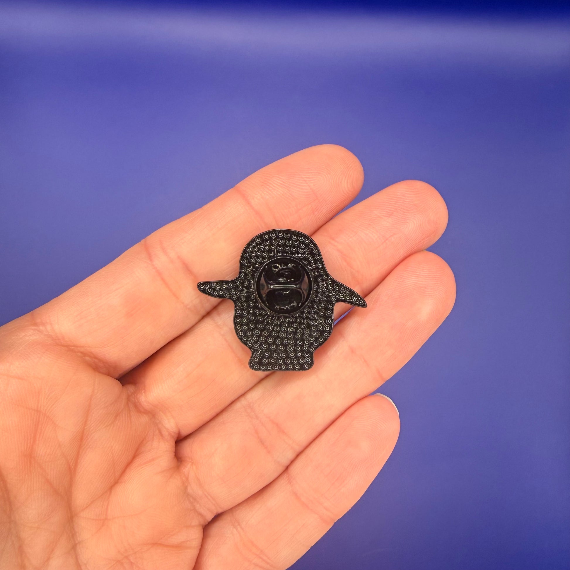 Back of the waddling penguin enamel pin showing the textured black metal back, held in hand