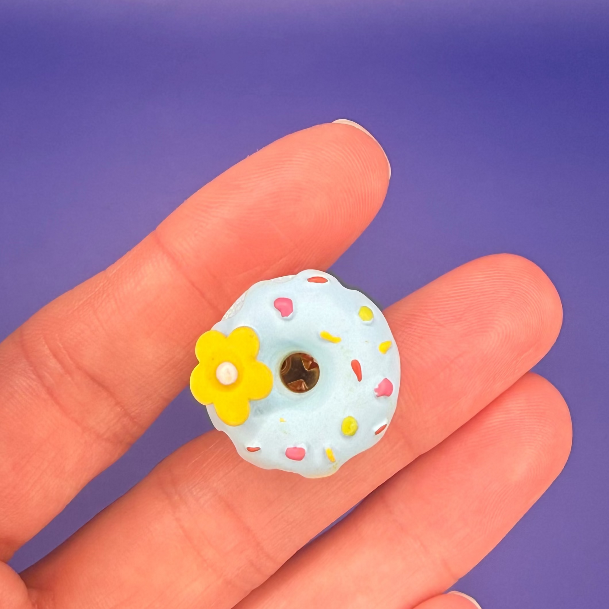 Powder blue donut enamel pin with colorful sprinkles and a yellow daisy, held in hand