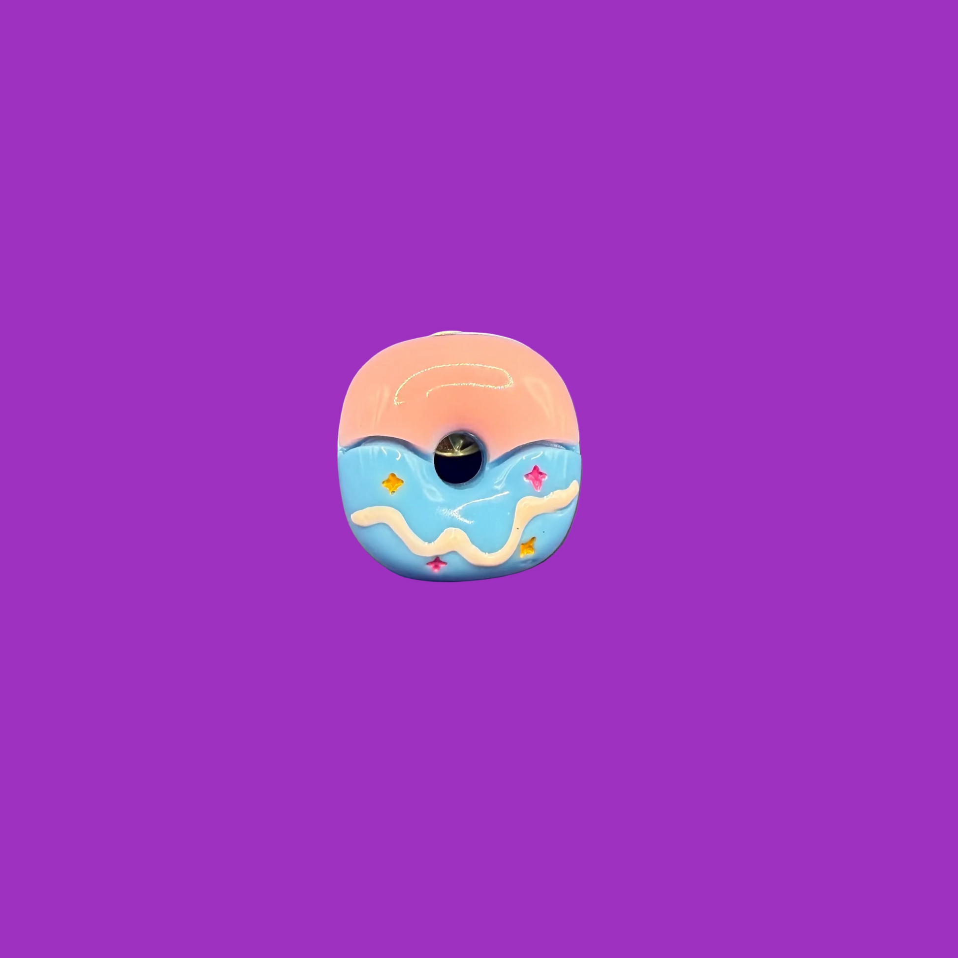 Pastel glazed donut enamel pin, half blush pink and half powder blue with a wavy white drip, on a purple background