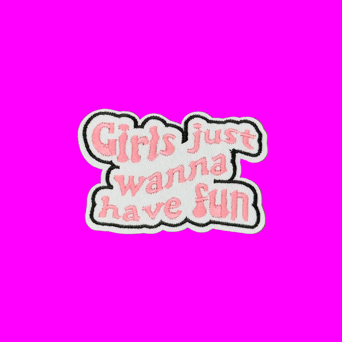 An embroidered patch with the phrase 'Girls just wanna have fun' in pink script on a white background with a black stitched border, shown on a solid brown surface.