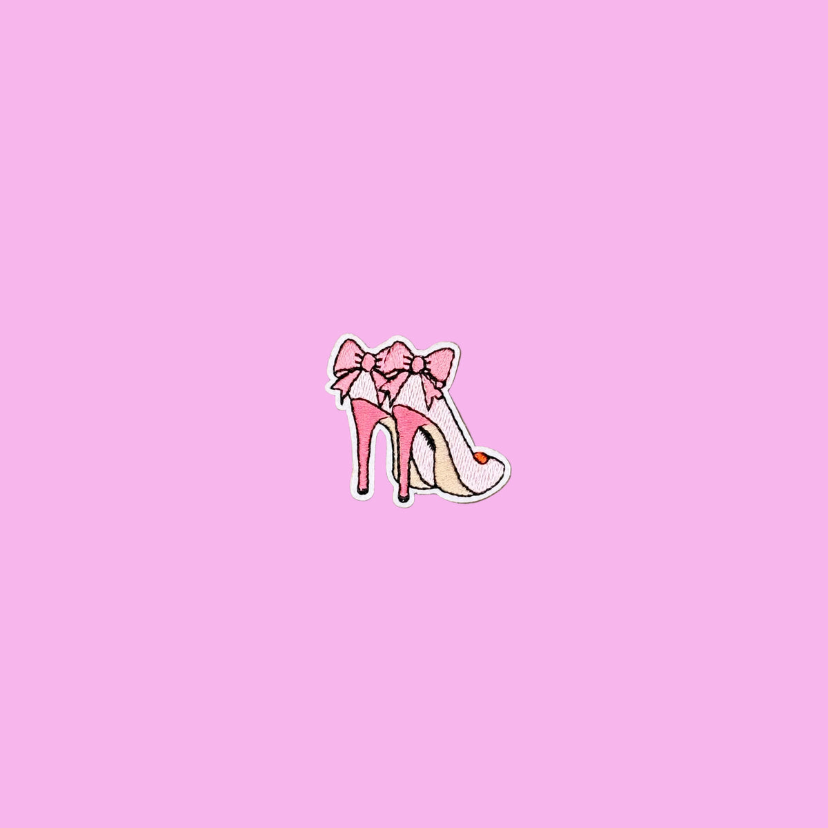 Small embroidered patch featuring a stylized pink high-heeled shoe with a bow, placed on a plain pink background.