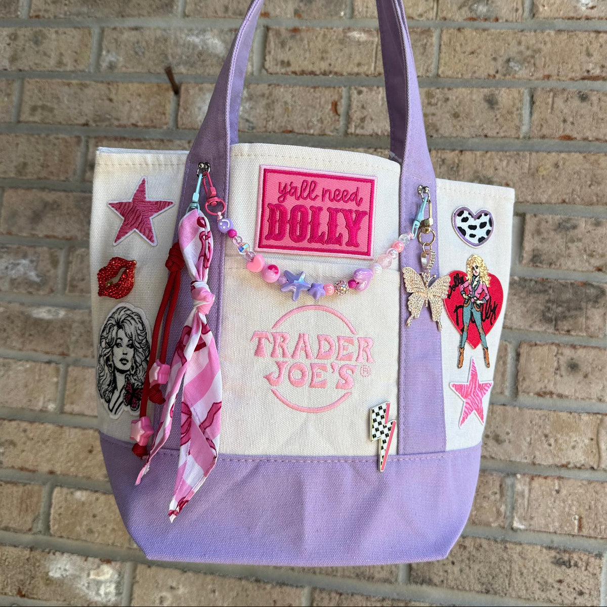 A lavender and cream Trader Joe's mini canvas tote decorated with pink and multicolor patches, charms and a beaded bag chain, hanging against a brick wall. Dolly Parton Pretty in Pastel mini tote by PrettyMUCH.