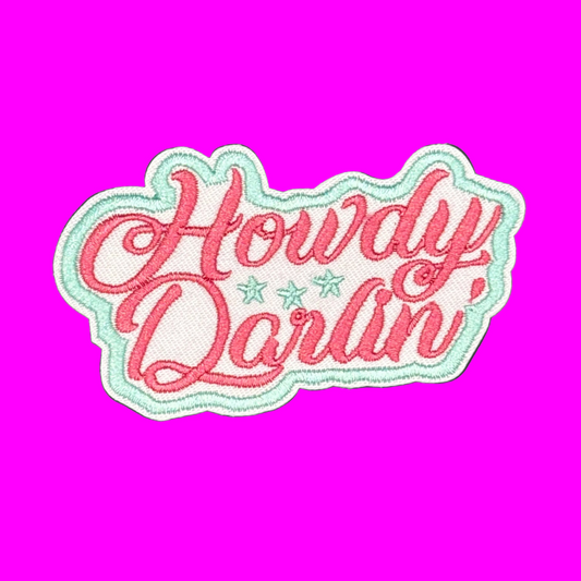 An embroidered patch with the phrase 'Howdy Darlin'' in pink cursive lettering, accented with two small mint green stars, all outlined with a white and mint green border, set against a solid purple background.