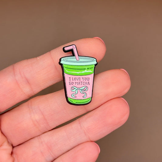 A small enamel pin shaped like a green drink cup with a pink label reading 'I LOVE YOU SO MATCHA' and a pink straw, held between fingers against a soft brown background.
