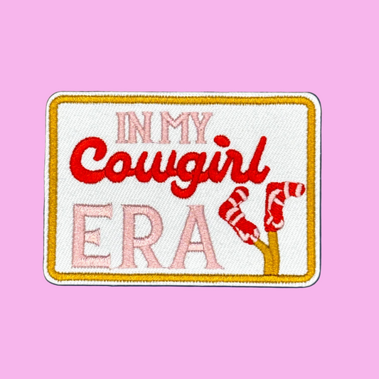 An embroidered rectangular patch with a white background and gold border, featuring the text 'IN MY Cowgirl ERA' in pink and red embroidery, plus a red-and-white striped leg design, shown on a solid blue background.