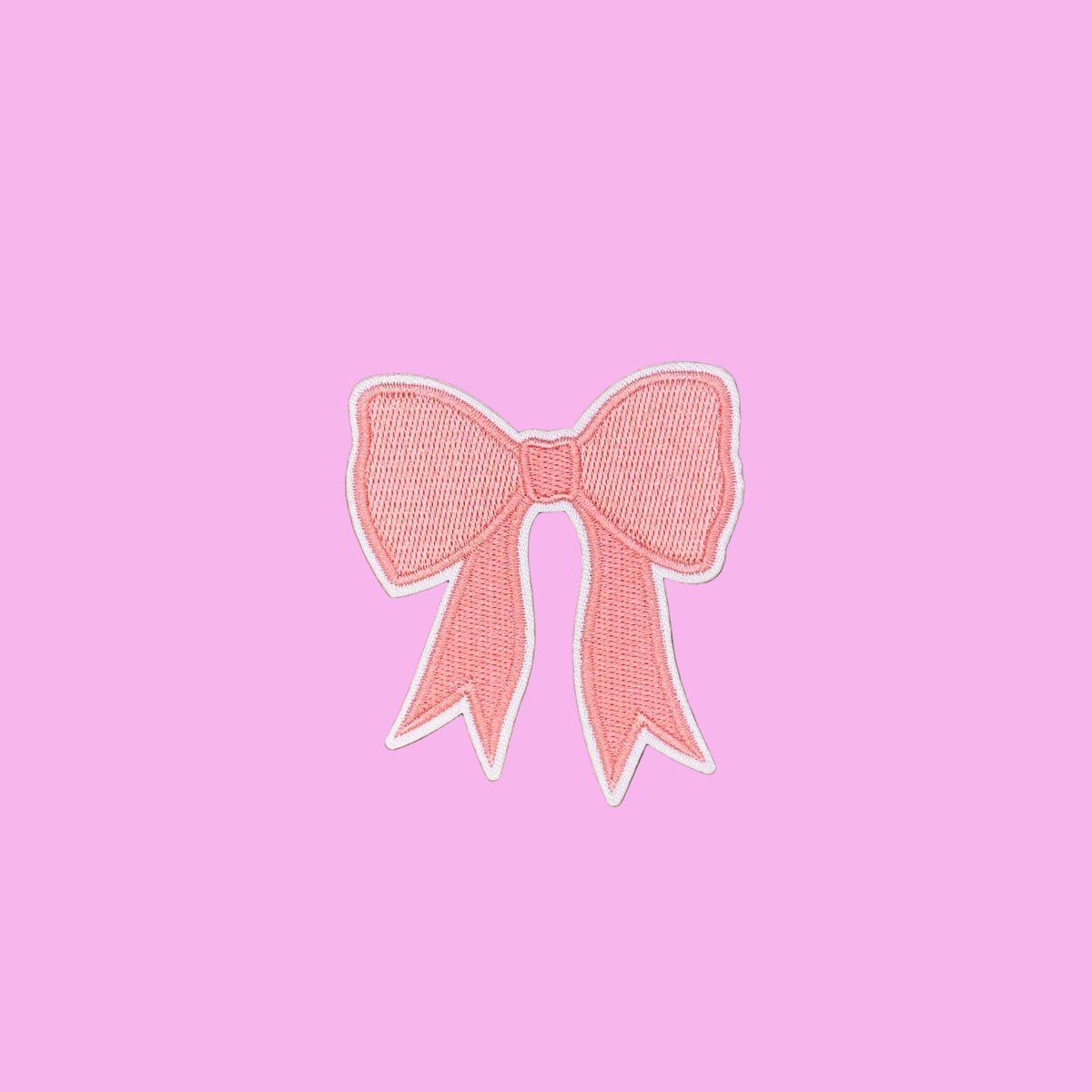A pink embroidered bow patch with a white border, displayed on a smooth brown background.