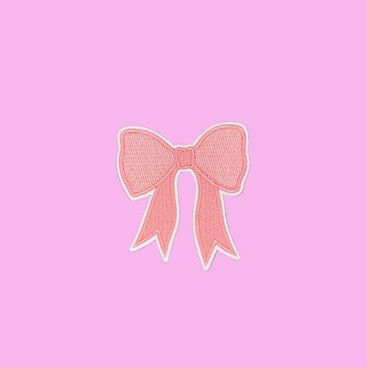 A pink embroidered bow patch with a white border, displayed on a smooth brown background.