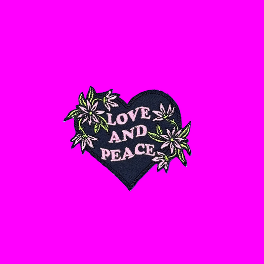 A heart-shaped black embroidered patch with the words 'LOVE AND PEACE' in pink, surrounded by pink and green flowers, shown on a solid blue background.