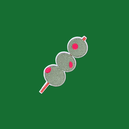 An embroidered patch of three green salami slices on a red skewer, displayed on a solid dusty rose background.