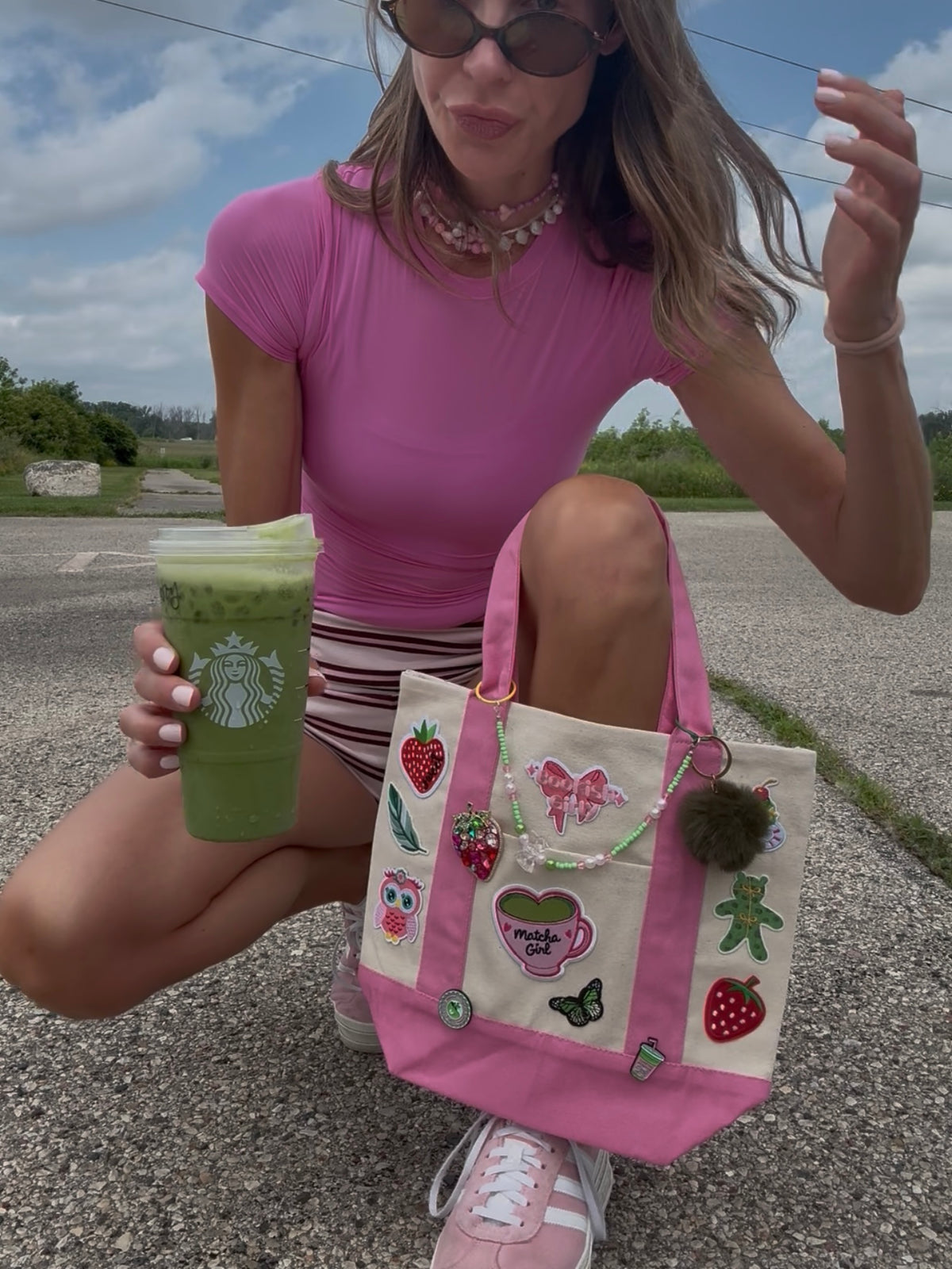 A woman crouches outdoors holding a green Starbucks drink and a pink and cream canvas tote bag decorated with strawberry, owl, and coffee cup patches.