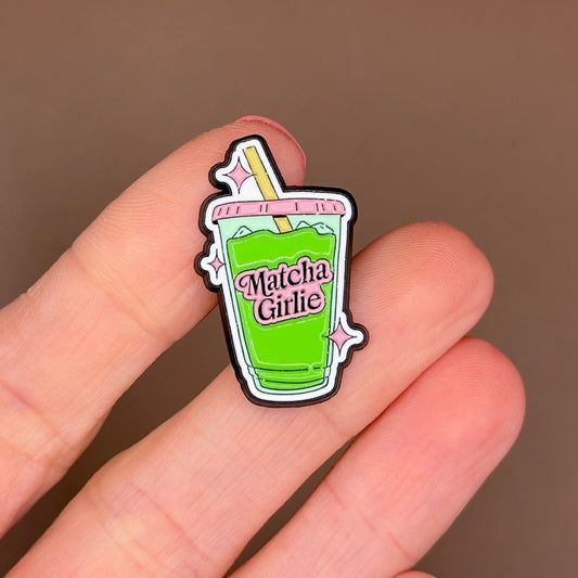 A small enamel pin shaped like a green matcha drink cup with the words 'Matcha Girlie' is held between fingers against a soft brown background.