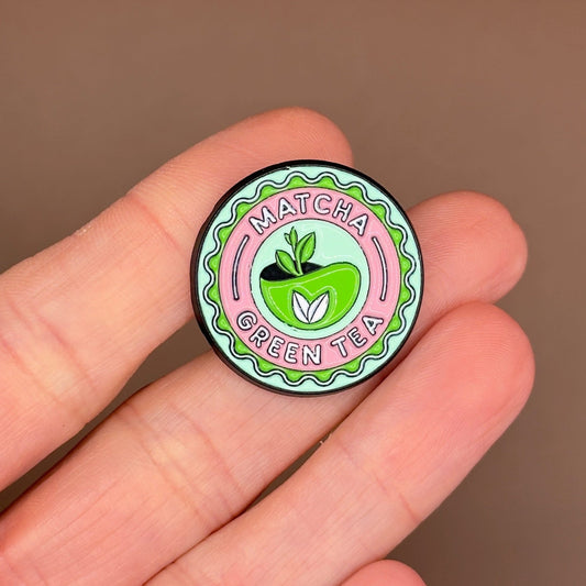 A small round enamel pin featuring a green apple with a matcha leaf and the words 'MATCHA GREEN TEA' on a pink and green patterned background, held between fingers against a warm brown backdrop.