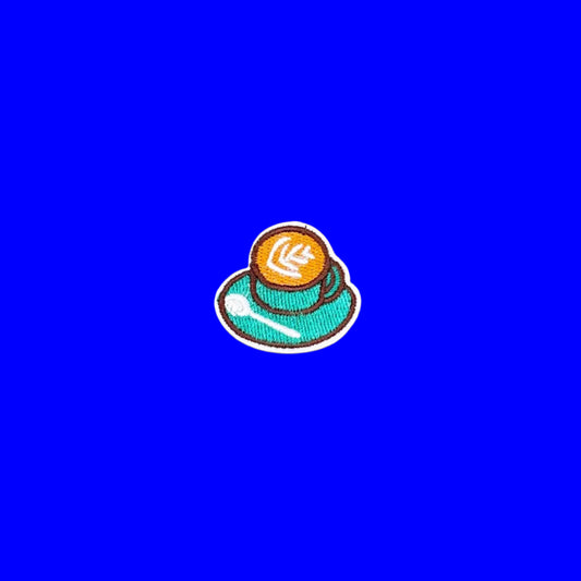 A small embroidered patch depicting a coffee cup with latte art, set against a solid blue textured background.
