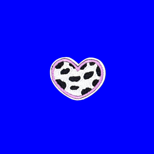 A heart-shaped embroidered patch featuring a black and white cow print pattern with a pink border, displayed on a solid blue background.