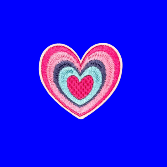 A heart-shaped embroidered patch with concentric layers of pink, magenta, navy, and light blue, finished with a white border, shown on a solid purple background.