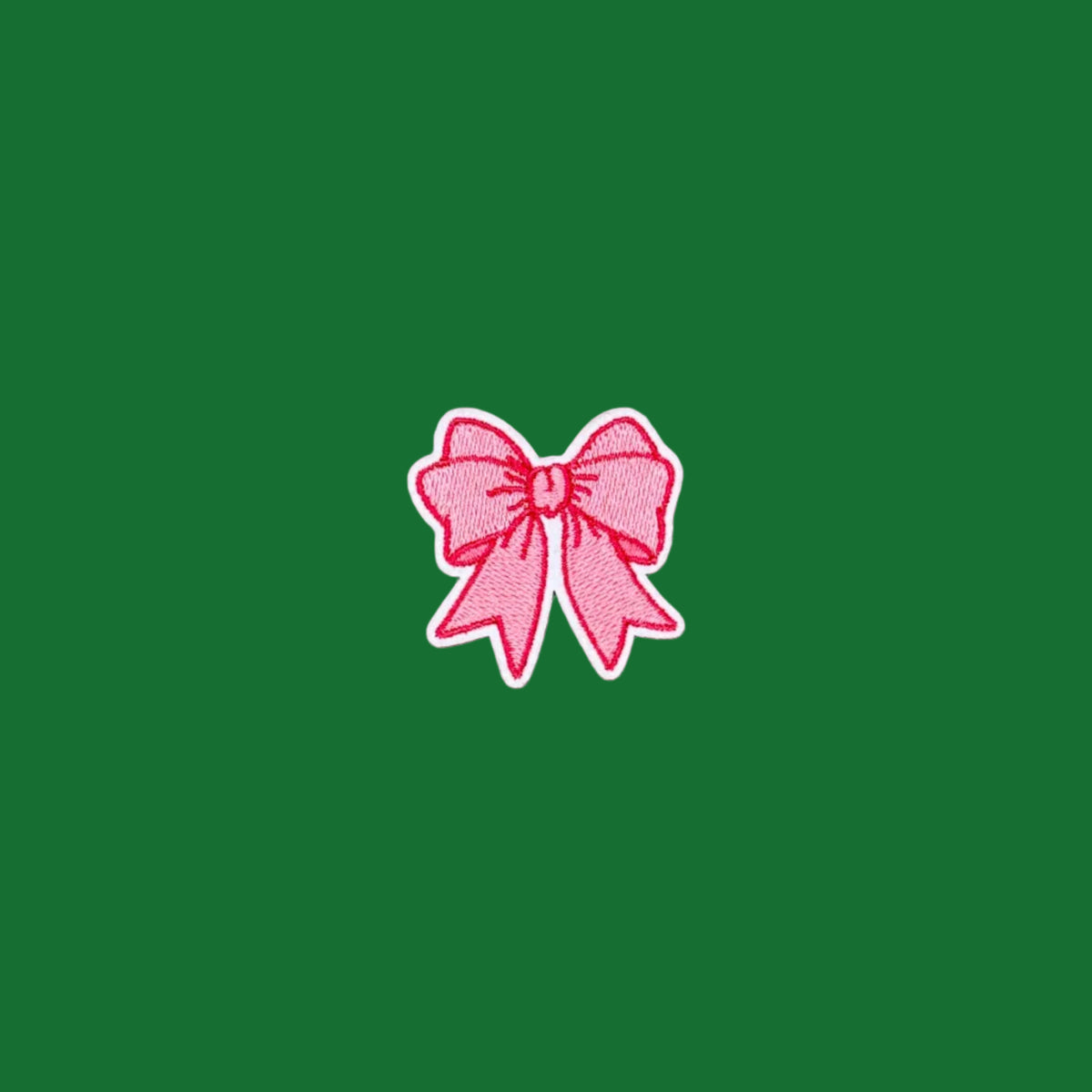 A small embroidered pink bow patch with a white border, centered on a solid green background.