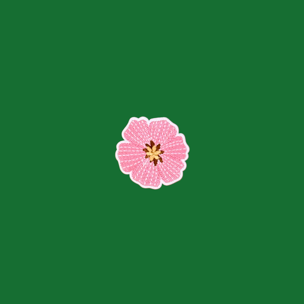 A small pink flower-shaped embroidered patch with a white border and a yellow-brown center, placed on a green background.
