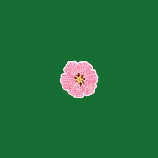 A small pink flower-shaped embroidered patch with a white border and a yellow-brown center, placed on a green background.