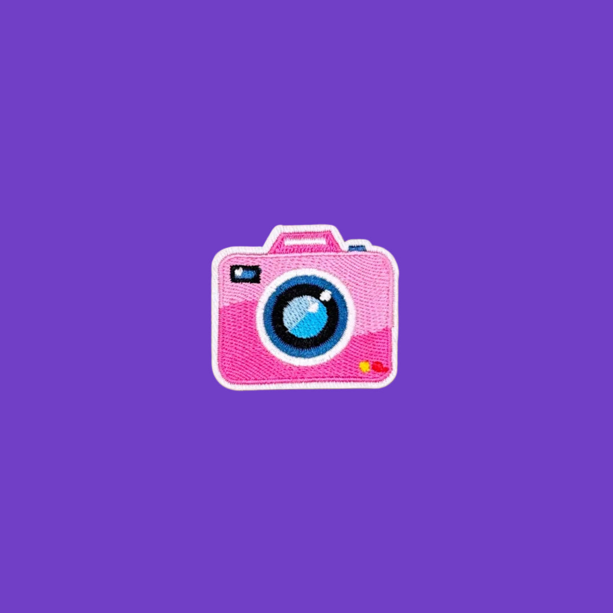 A pink embroidered patch shaped like a retro camera, featuring a blue and white lens and a small yellow and red detail, centered on a solid purple background.