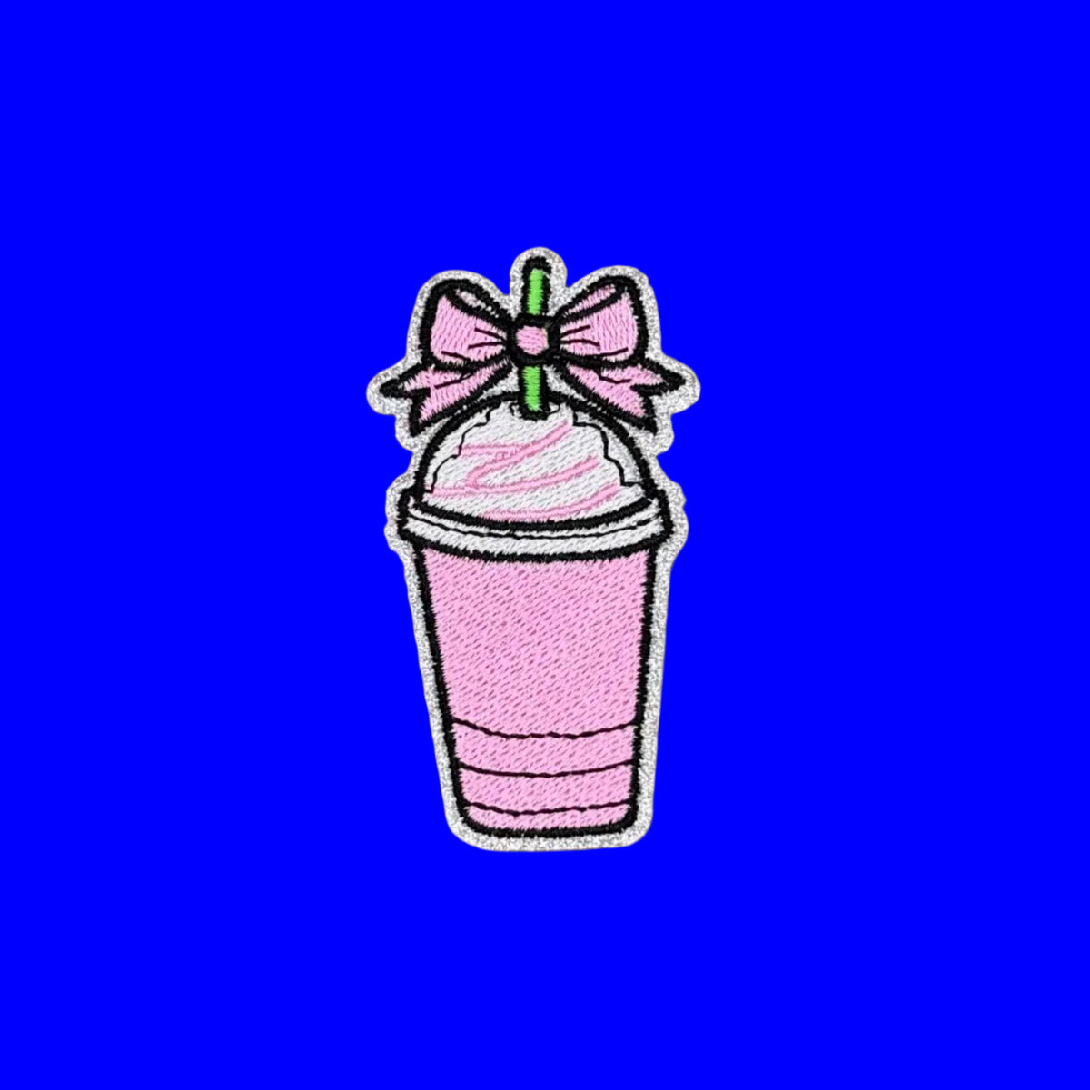 A pink embroidered patch shaped like a cup of milkshake or smoothie with whipped topping, a pink bow, and a green straw, shown on a blue background.