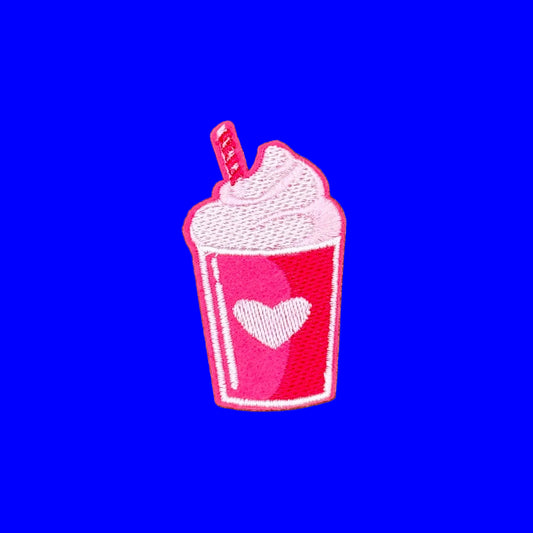 An embroidered patch shaped like a pink drink cup with white whipped cream, a pink heart on the front, and a small striped straw, shown on a plain brown background.