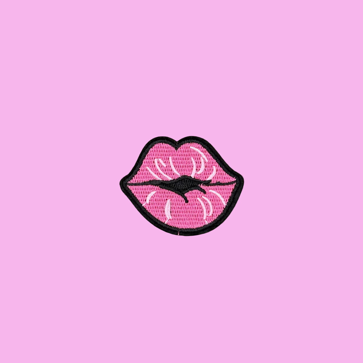 A pink embroidered patch shaped like lips with white highlights and a black border, placed on a smooth tan surface.
