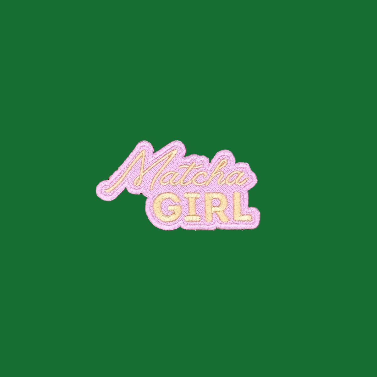 A die-cut embroidered patch reading 'Matcha GIRL' in pale pink and gold, placed on a smooth green background.