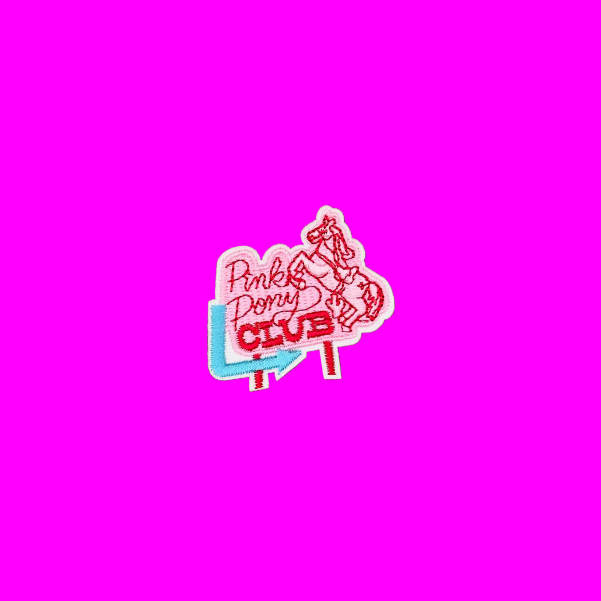 A small embroidered patch reading 'Pink Pony CLUB' in pink and red lettering with a light blue shape behind it, placed on a plain brown background.