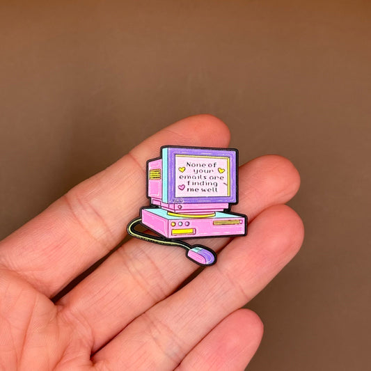 A small enamel pin shaped like a retro computer monitor and keyboard, held on a person's fingers against a plain brown background.