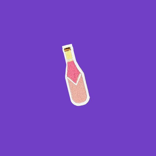 A small embroidered patch shaped like a champagne bottle, featuring pink and peach shades with a gold neck and white border, placed on a solid purple background.