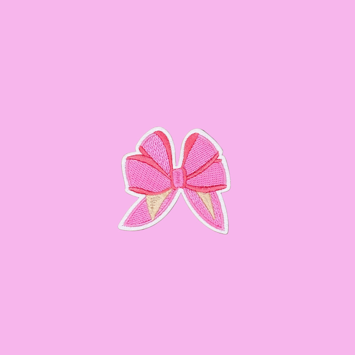 A pink embroidered bow patch with a white border, displayed on a smooth brown background.