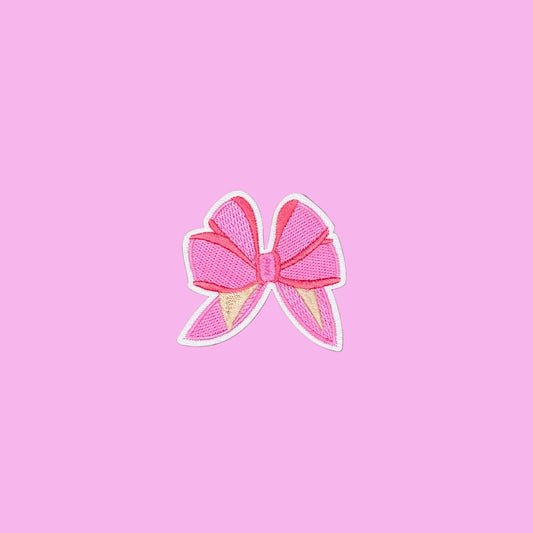 A pink embroidered bow patch with a white border, displayed on a smooth brown background.