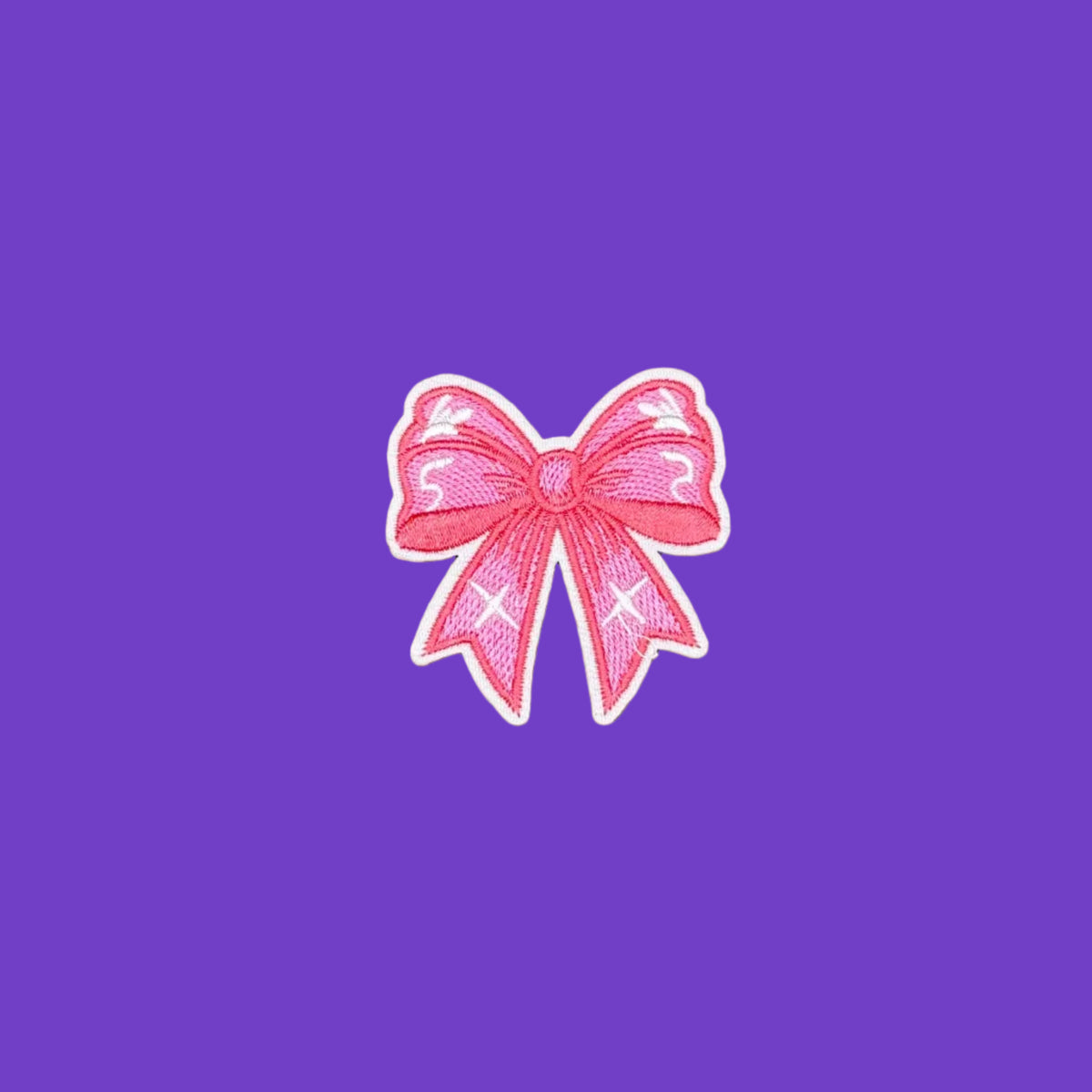 A pink embroidered bow patch with white embroidered details and a white border, centered on a brown textured background.