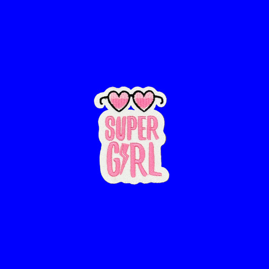 A die-cut sticker featuring the words 'SUPER GIRL' in pink bubble letters on a white background, topped with two pink hearts outlined in black, shown on a plain brown surface.