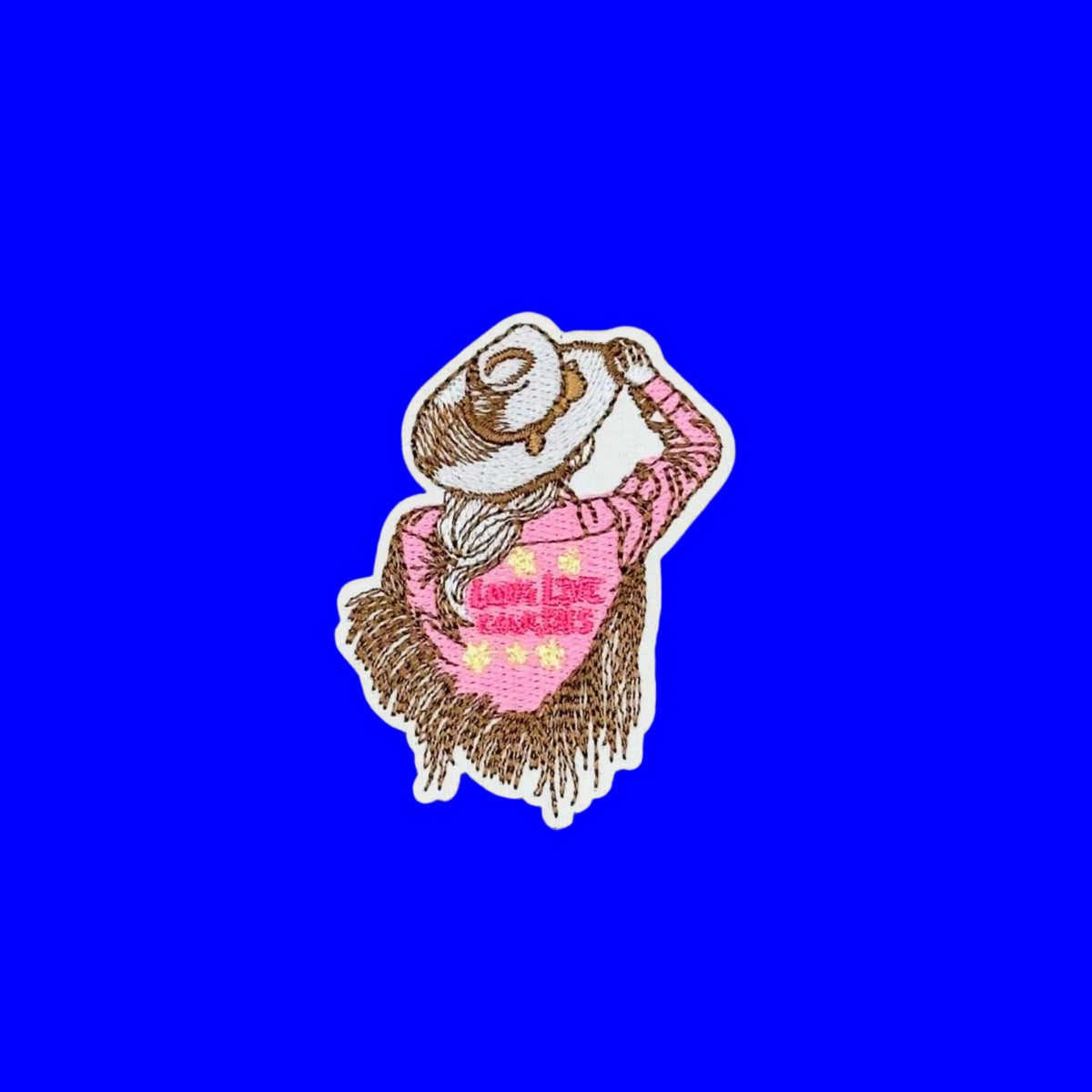 A small embroidered patch featuring a cowgirl figure in a pink shirt with yellow stars and brown hair, outlined in white, placed on a plain blue background.