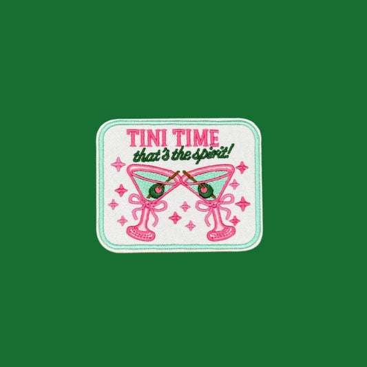 A rectangular embroidered patch with rounded corners featuring the text 'TINI TIME that's the spirit!' above two crossed pink martini glasses with green olives, set against a solid brown background.